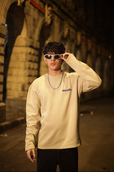 Man wearing a beige sweatshirt with a logo, sunglasses, and a chain in a dimly lit tunnel.